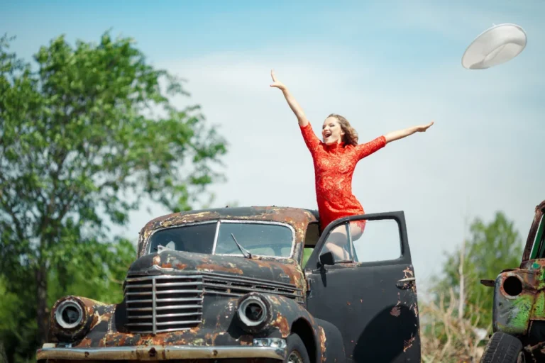 Femme en robe rouge sur une vieille voiture rouillée, souriant bras levés.