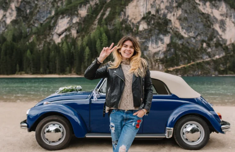 Femme souriante devant un cabriolet bleu près d'un lac et des montagnes.