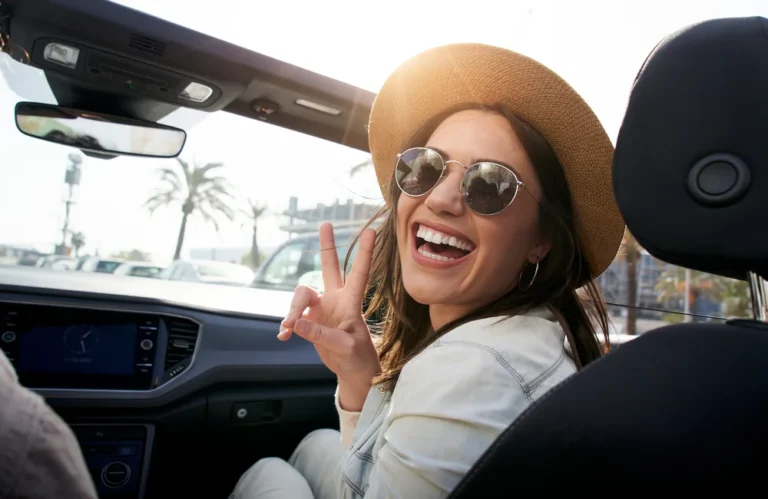 Femme souriante en chapeau, assise dans une décapotable, faisant un signe de paix.